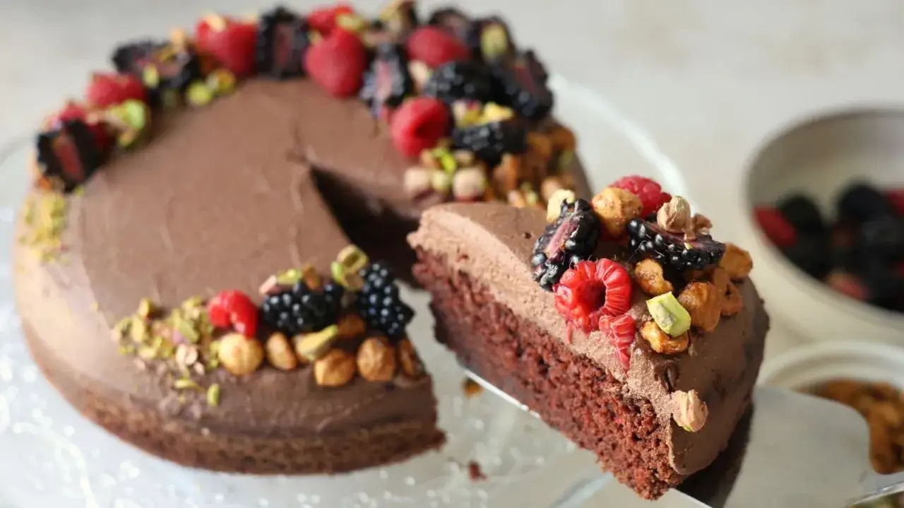 En saftig protein kladdkaka toppad med chokladfrosting, f&auml;rska b&auml;r och n&ouml;tter. En bit lyfts fram.