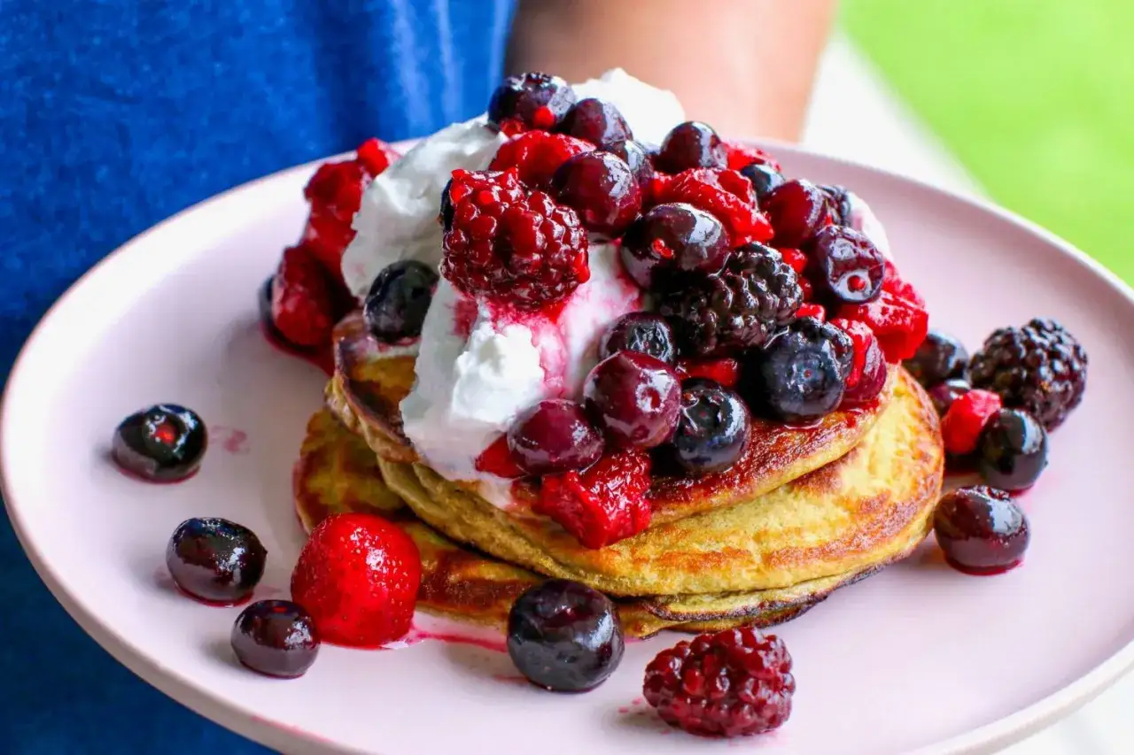 En hög med fluffiga proteinpannkakor toppade med vispgrädde och färska bär. Ett perfekt recept på proteinpannkakor för en hälsosam frukost.