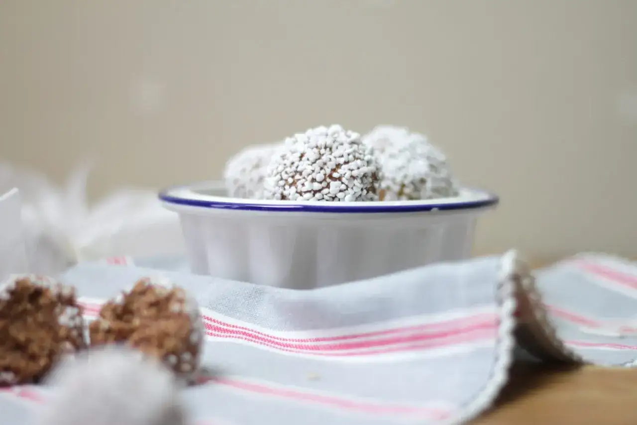 Små, runda havrebollar rullade i kokosflingor ligger i en vit skål. Ett enkelt havrebollar recept för fika.