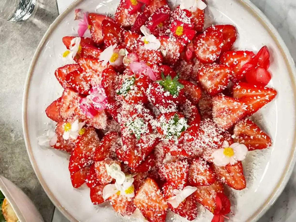 En vacker tallrik med skivade jordgubbar, strösslade med kokosflingor och dekorerade med små blommor. Ett bevis på att jordgubbar nyttigt kan vara både gott och vackert.