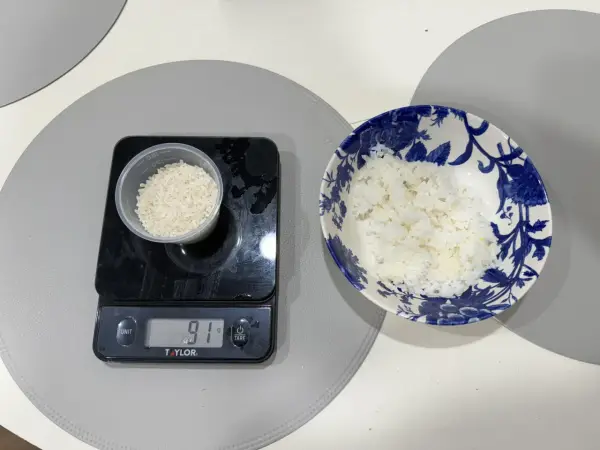 En digitalvåg visar 91 gram ris i en liten behållare. Bredvid står en skål med mer ris, redo att ätas. Perfekt för att hålla koll på råris kalorier.