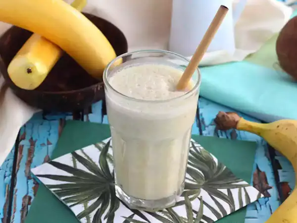 En krämig banan milkshake med en bambustrå i ett glas, omgiven av bananer och en kokosnöt.